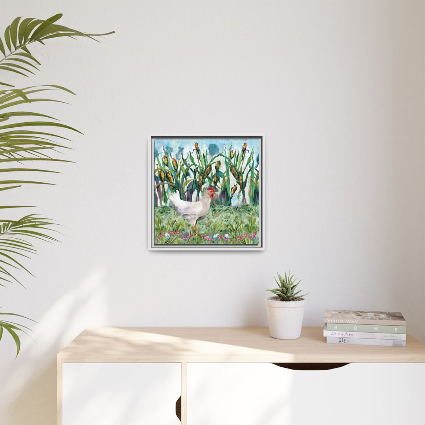 Framed artwork of chickens in a field on a wall above a desk with a plant and books.