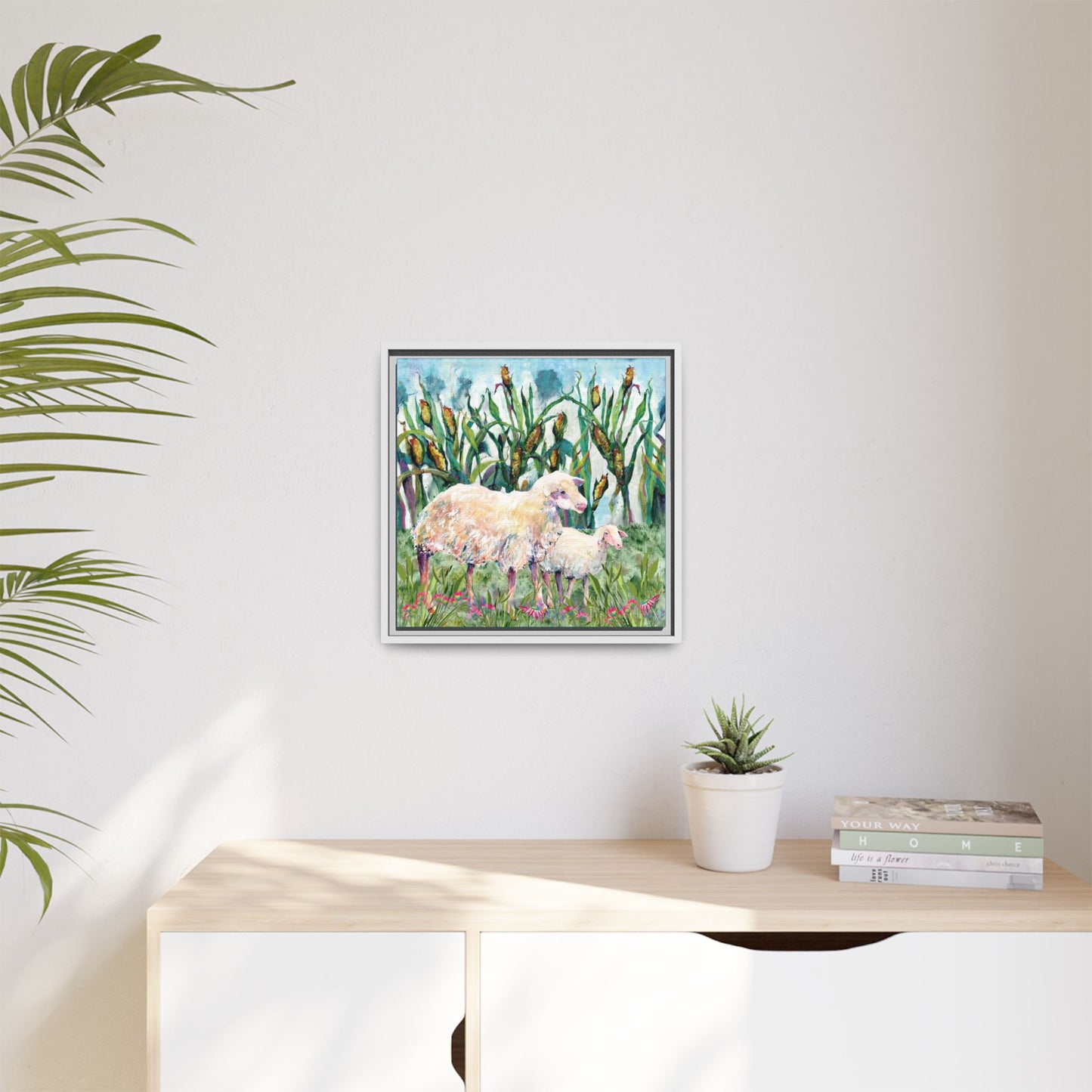 Framed artwork of sheep in a field on a wall above a desk with a plant and books. White frame