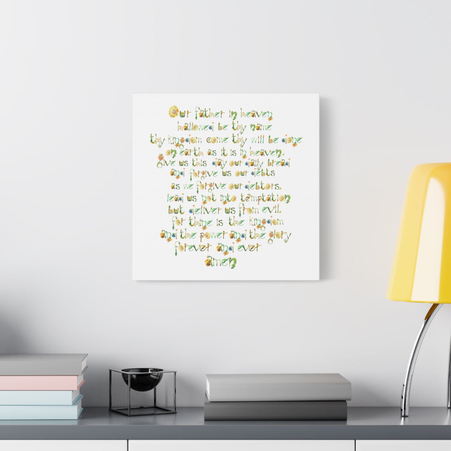 Canvas print of The Lords Prayer in hand-drawn floral font, on a white wall above a desk with books and a lamp.