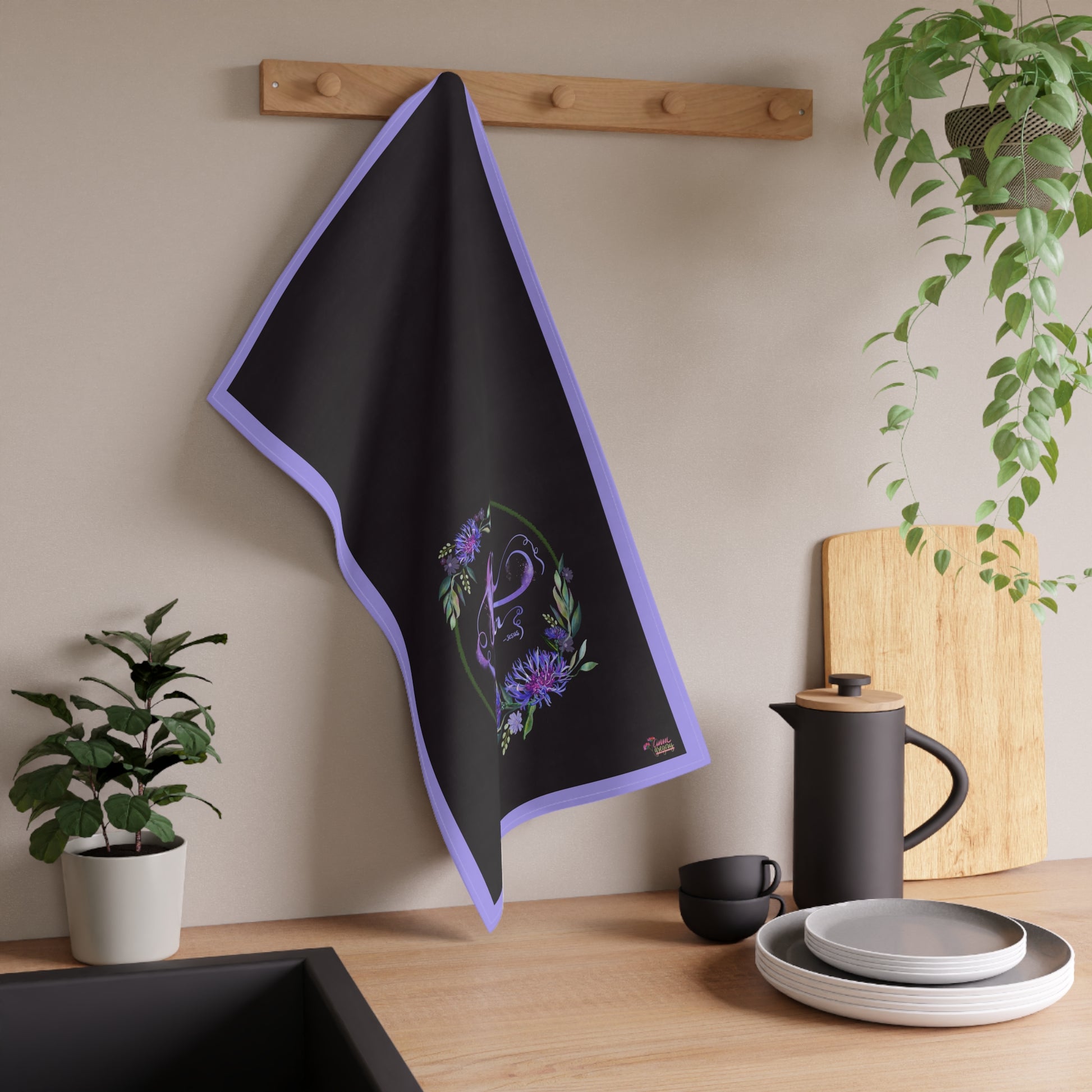 Black kitchen tea towel with purple trim and bible verse text Loe One Another hanging on a wooden rack in a kitchen setting.