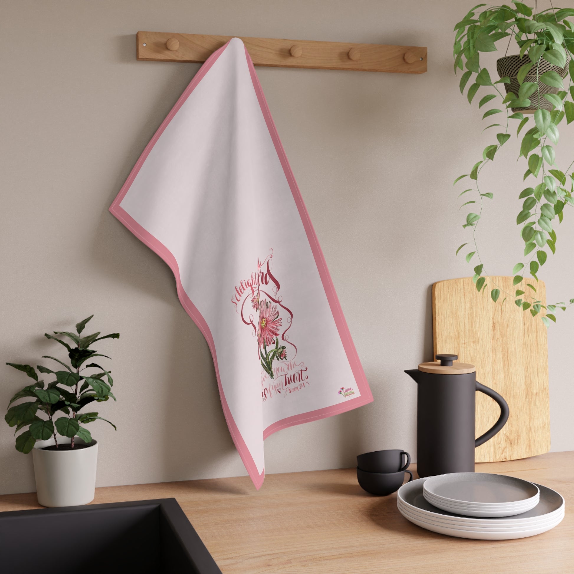 Kitchen towel with floral design with Bible Verse hanging on a wooden rack, next to a plant and kitchen items.