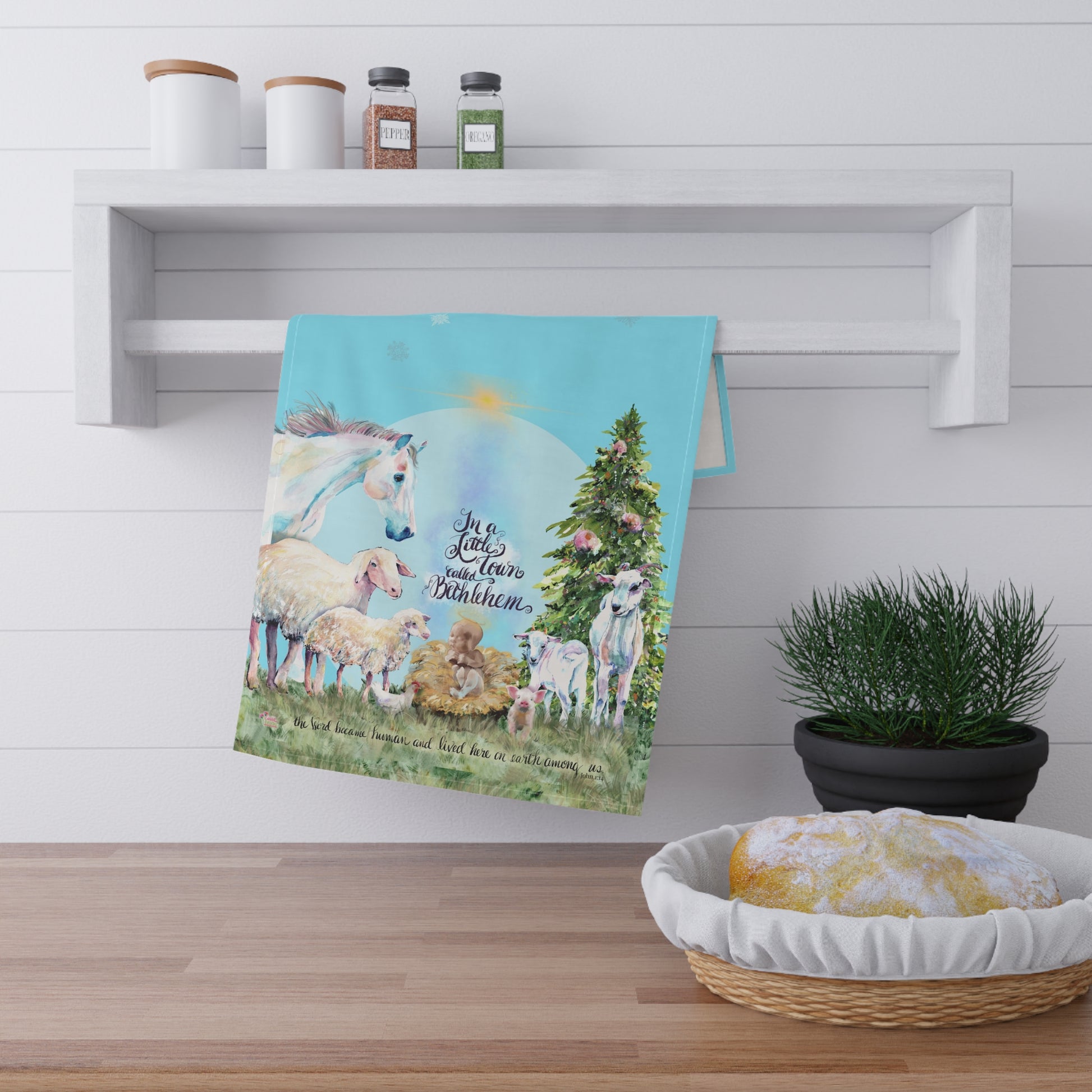 Kitchen Towel with Christmas scene of sheep and a lamb owith text in a little town called Bethlehem on a wall above a wooden shelf with a plant and basket.