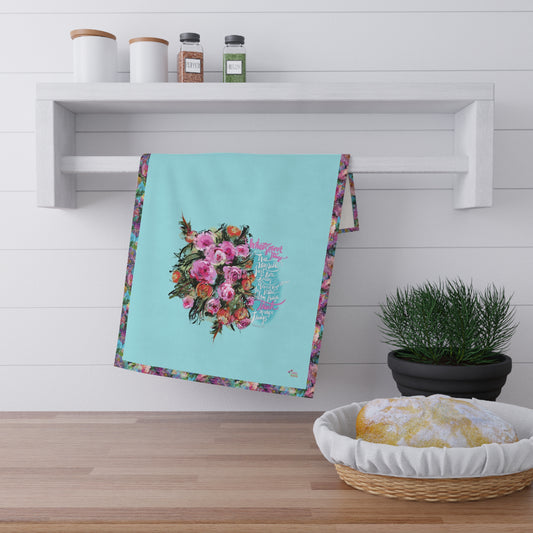 Floral kitchen tea towel with Bible verse "Whatsoever things"hanging on a wooden shelf with a plant and basket in the foreground.