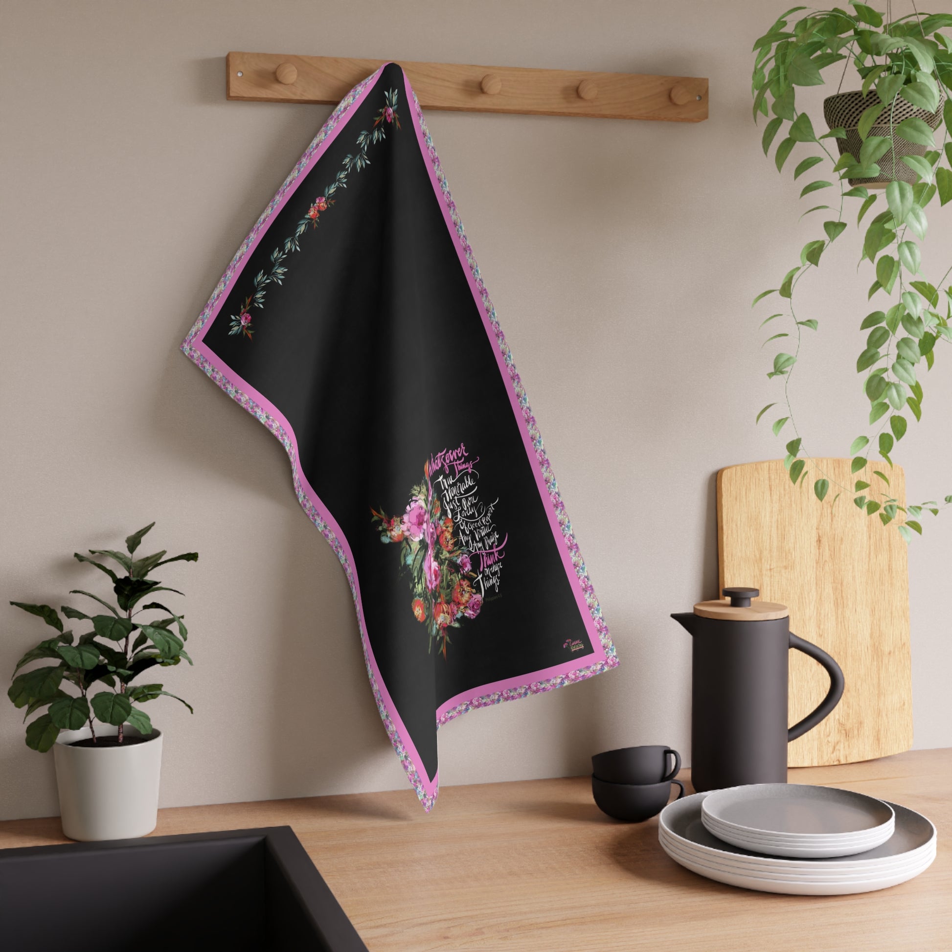 Black kitchen tea towel with floral design and Bible verse on a wooden hook against a neutral wall with kitchen items below.