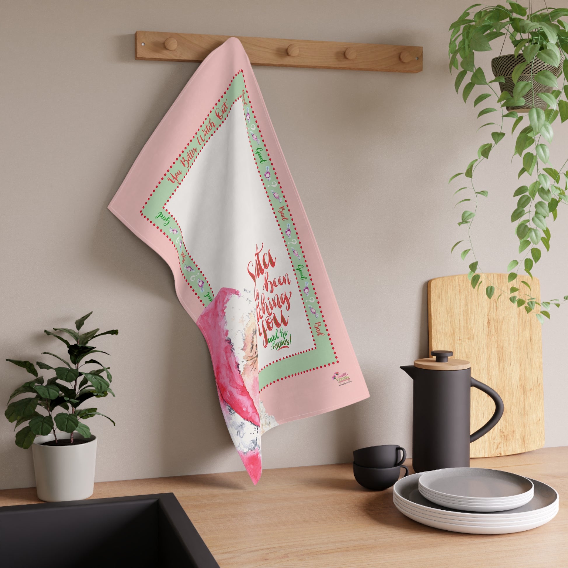 Decorative tea towel with Christmas Santa design and text hanging on a wooden rack in a kitchen setting.