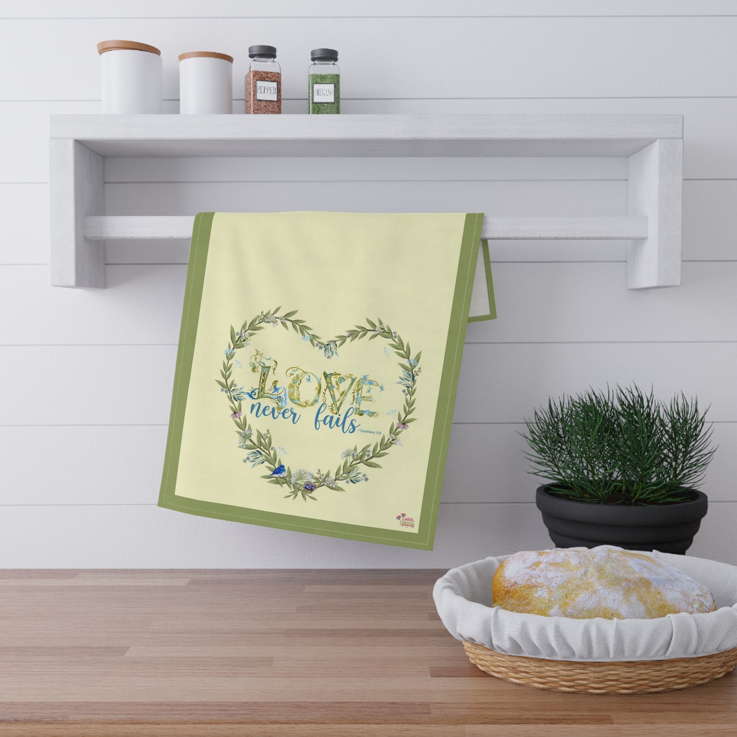 Decorative litchen towel with 'Love never fails' text on a kitchen shelf with a plant and bread basket.