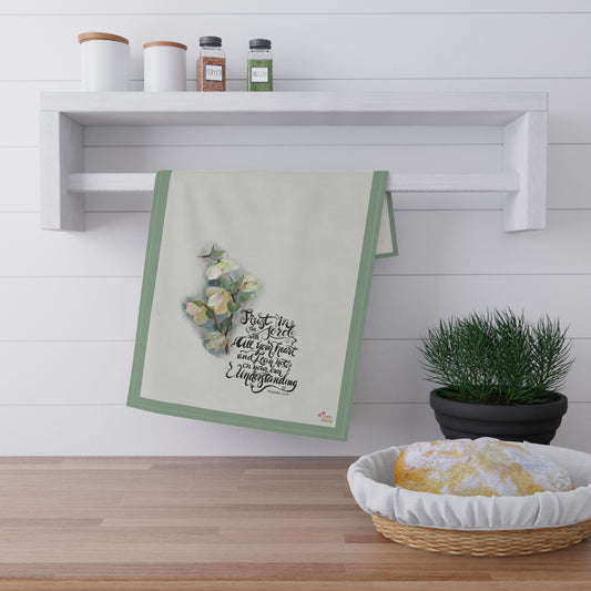 Kitchen towel with Bible verse calligraphy "Trust in the Lord " and flowers hanging on a wooden shelf with a plant and bread basket below.