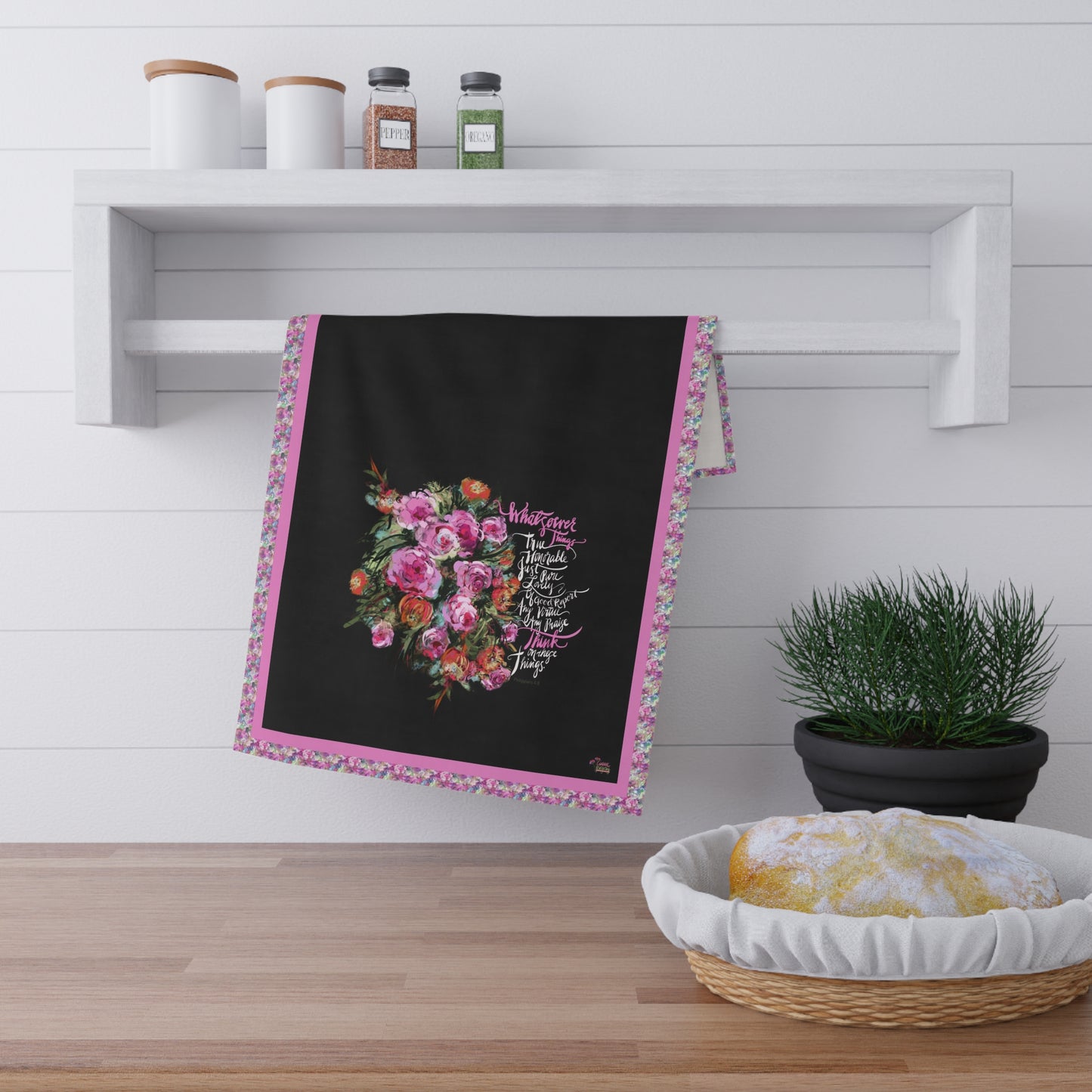 Decorative kitchen tea towel with floral design and Bible verse "Whatsoever things" on a wooden surface with a plant and basket.