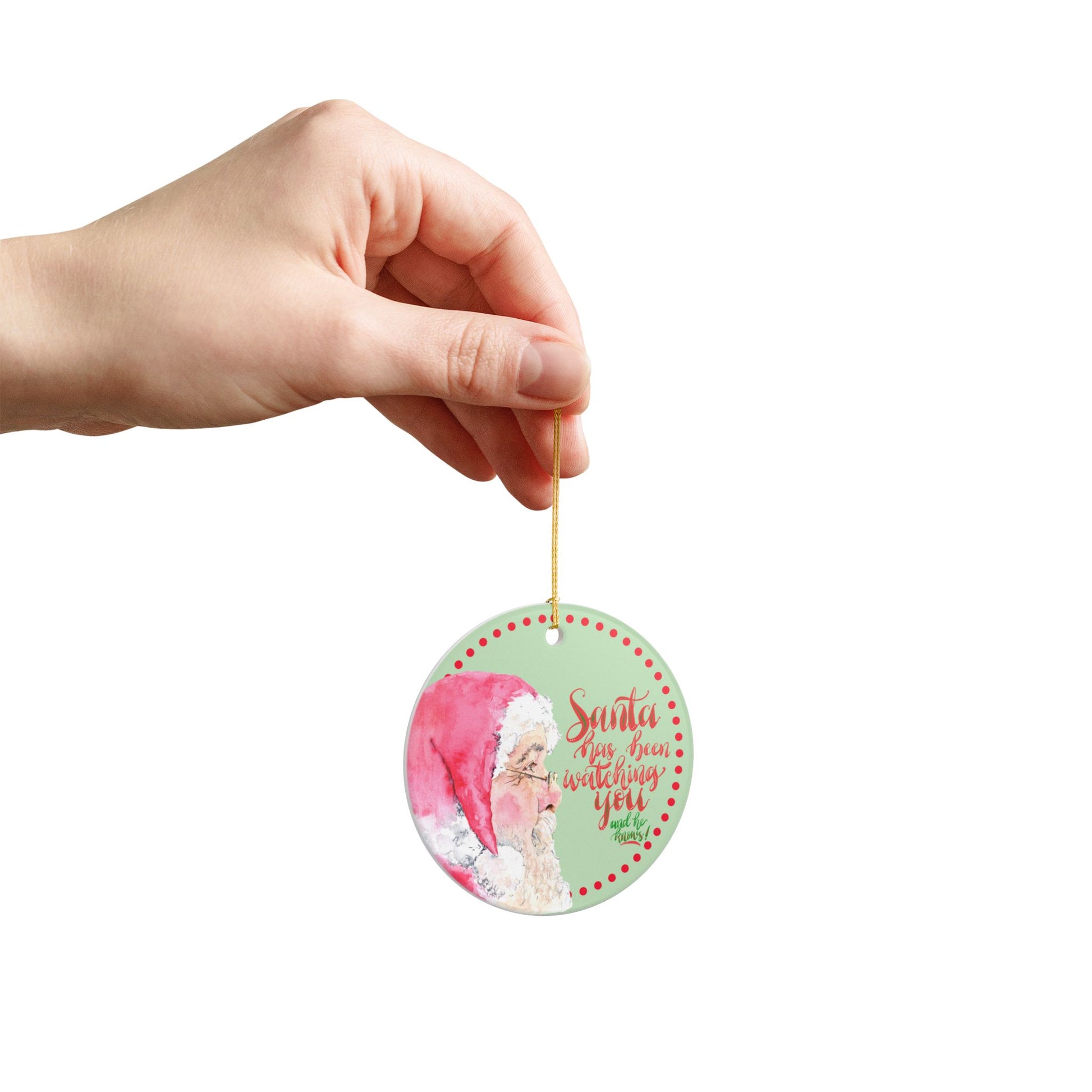 Hand holding a round decorative Christmas ornament with a Santa Claus design and text on a mint green background