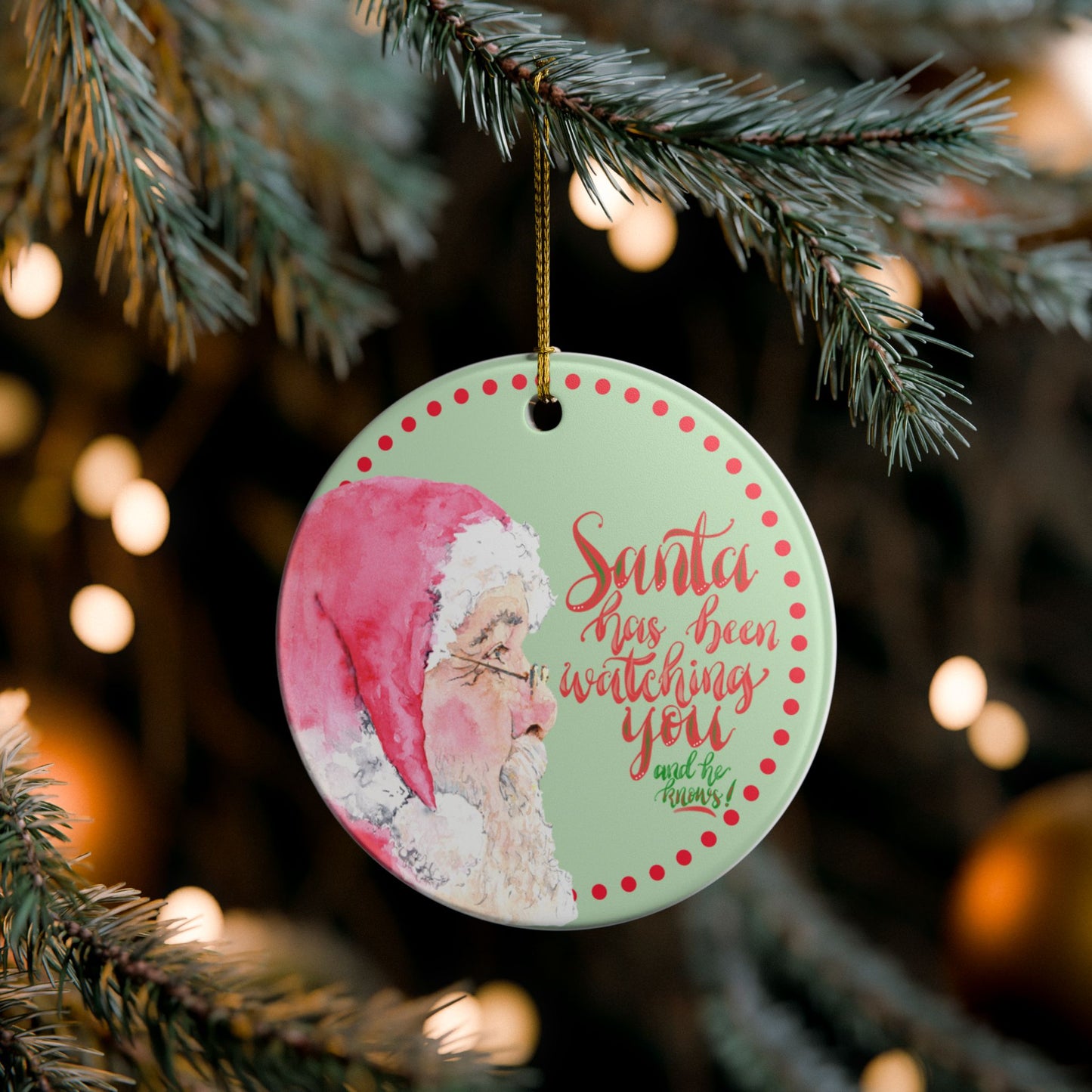 Christmas ornament with Santa Claus and text Santa has been watching you and he knows on a tree branch with blurred lights in the background