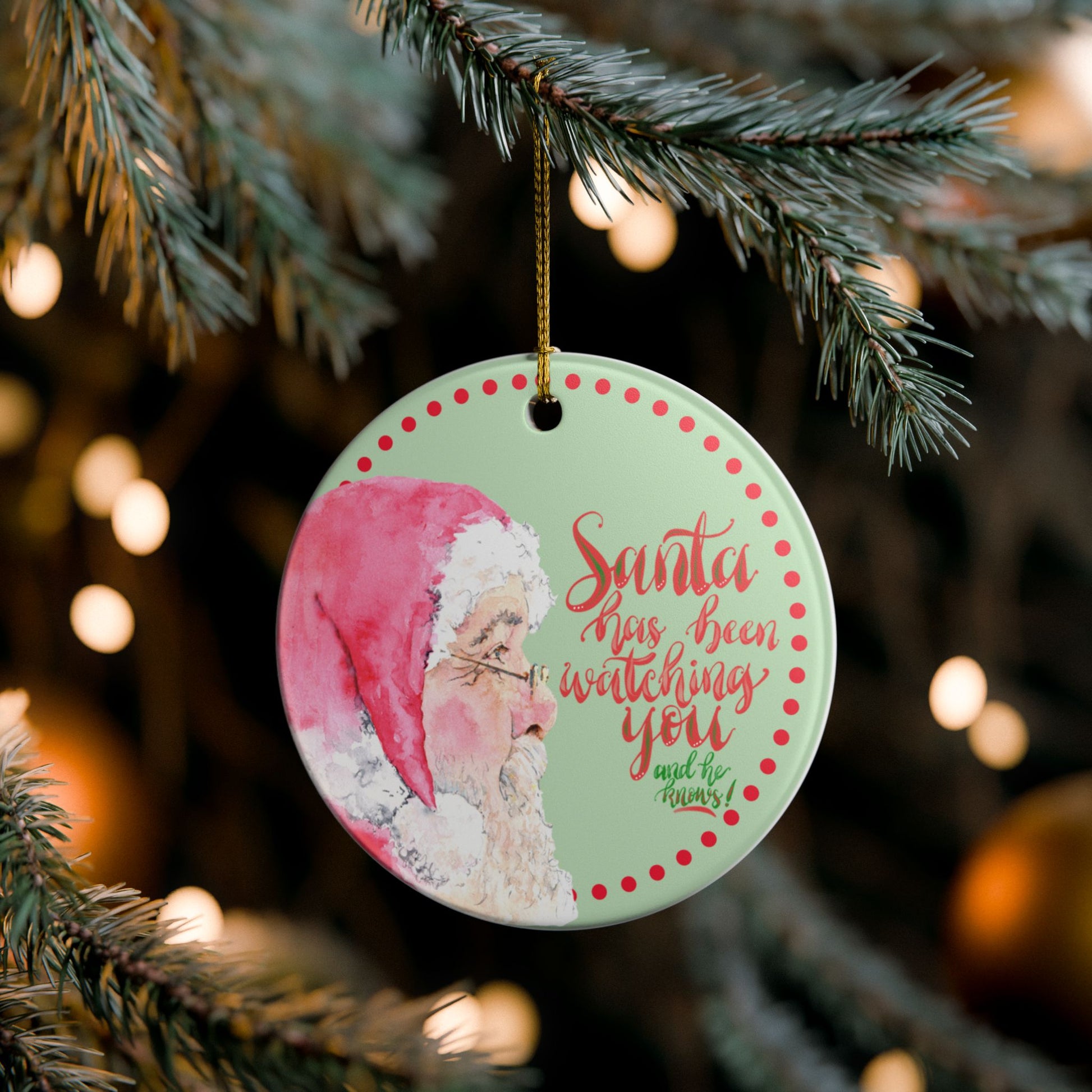 Christmas ornament with Santa Claus and text Santa has been watching you and he knows on a tree branch with blurred lights in the background