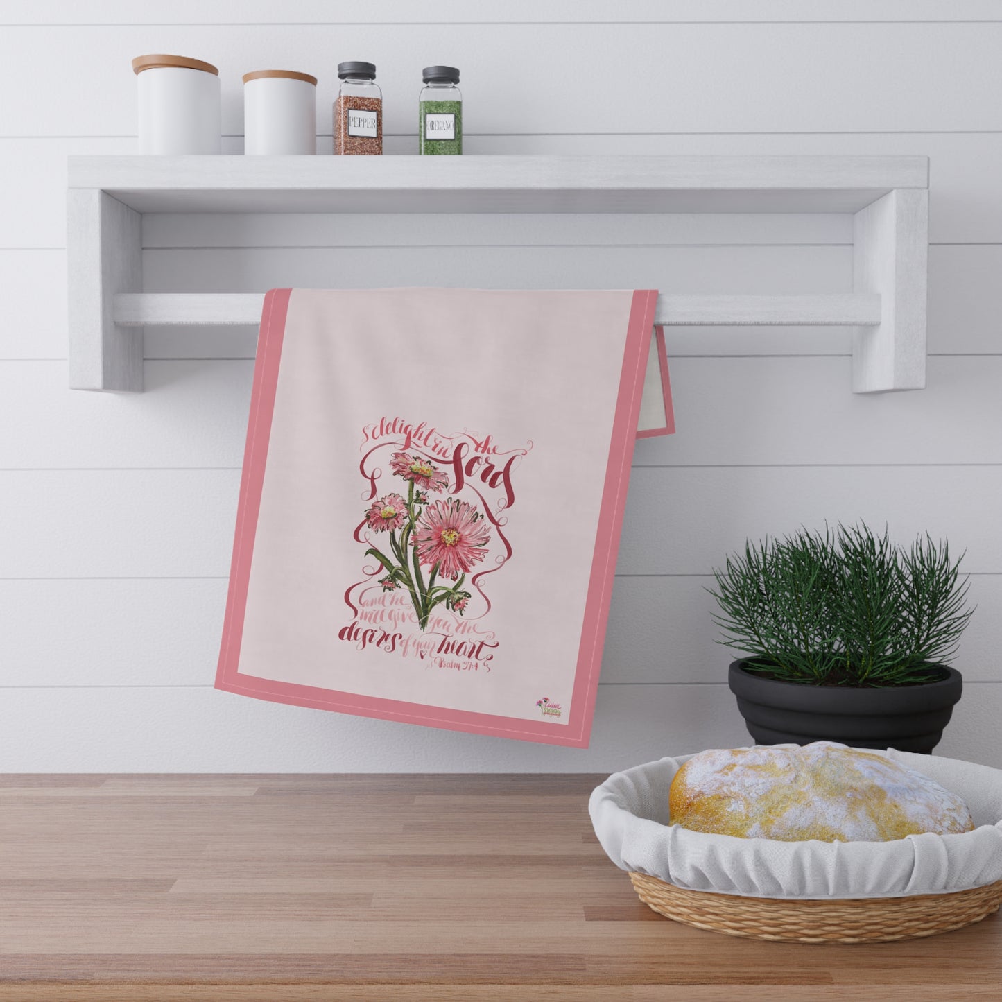 Decorative kitchen towel with floral design and Bible verse "Delight i the Lord"  hanging on a white wall above a wooden shelf.