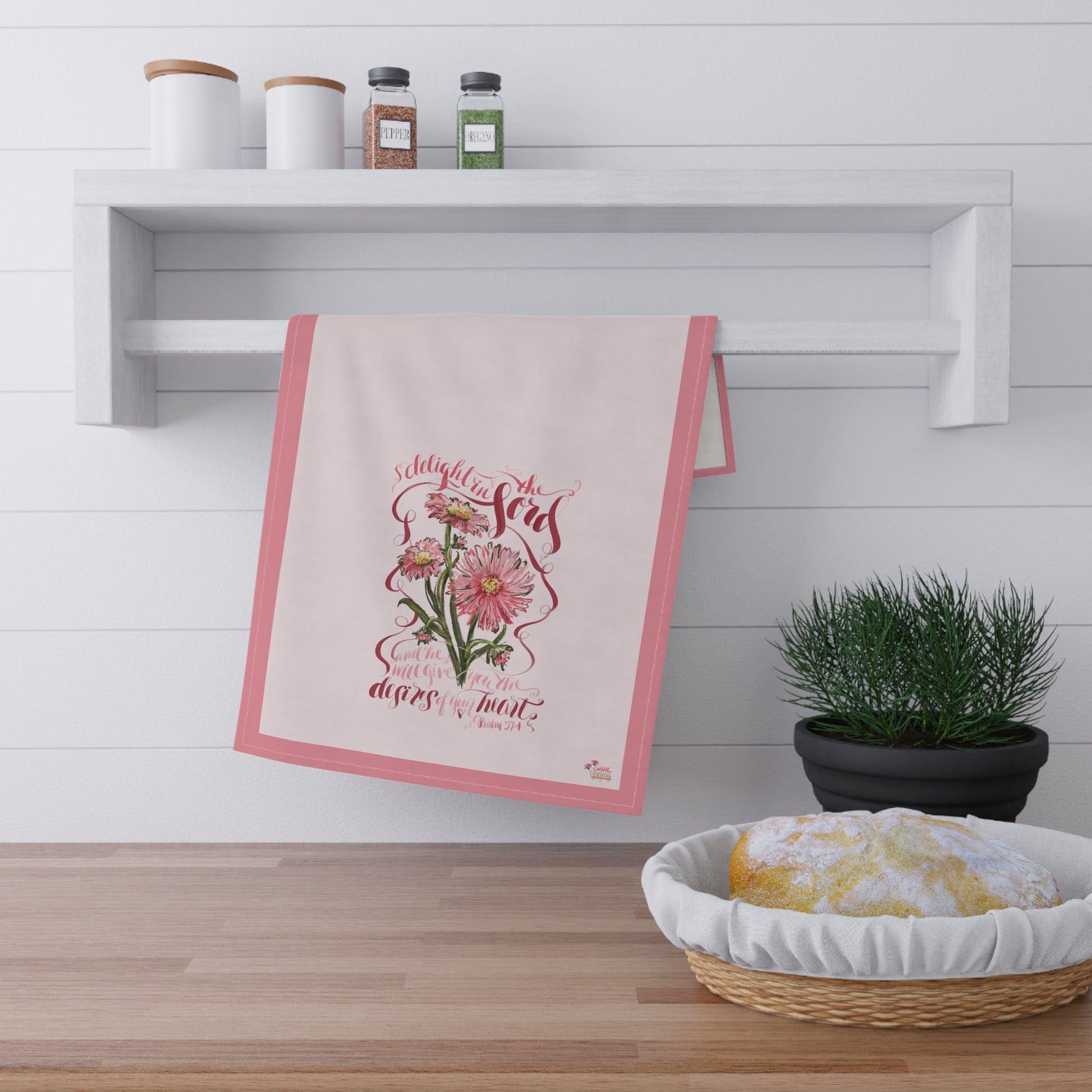 Decorative kitchen towel with floral design and Bible verse "Delight i the Lord"  hanging on a white wall above a wooden shelf.