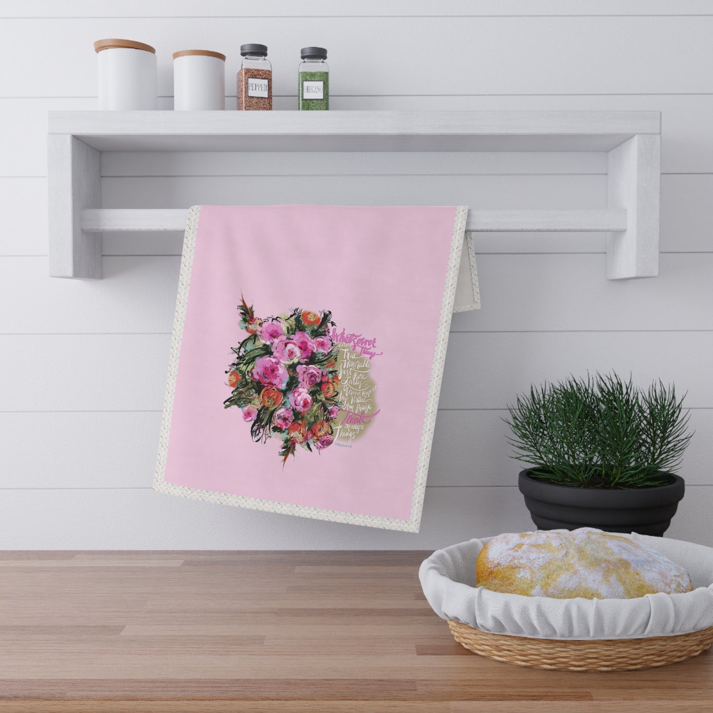 Pink tea towel with Bible verse "Whatsoever things" and floral design hanging on a white shelf in a kitchen setting.