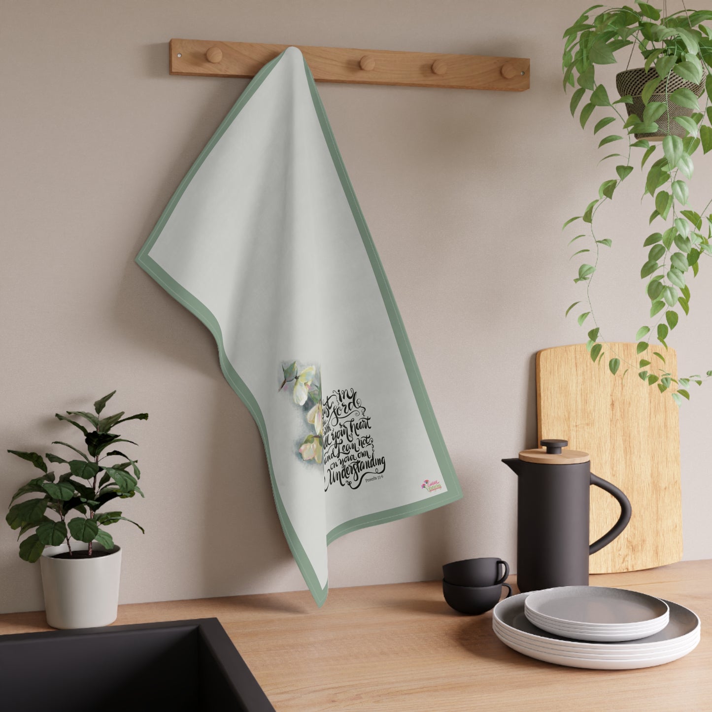 Kitchen towel with Bible verse design hanging on a wooden rack, next to a plant and kitchen items.