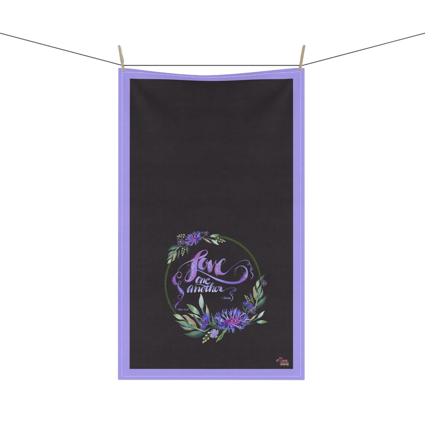Black tea towel with text Bible Verse Love One Another purple border and floral design hanging on a line.