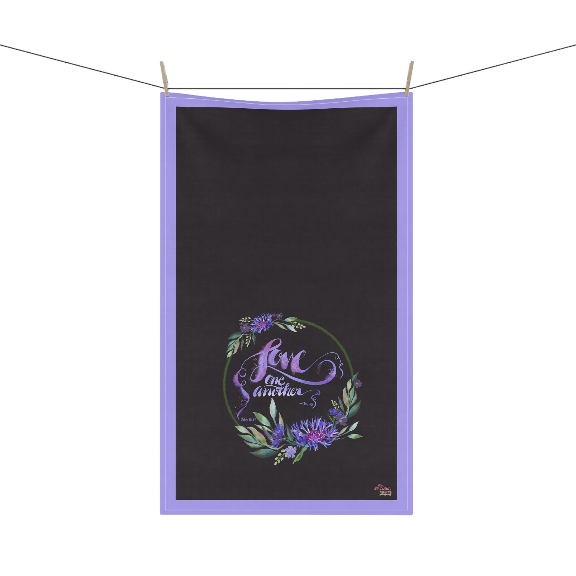 Black tea towel with text Bible Verse Love One Another purple border and floral design hanging on a line.