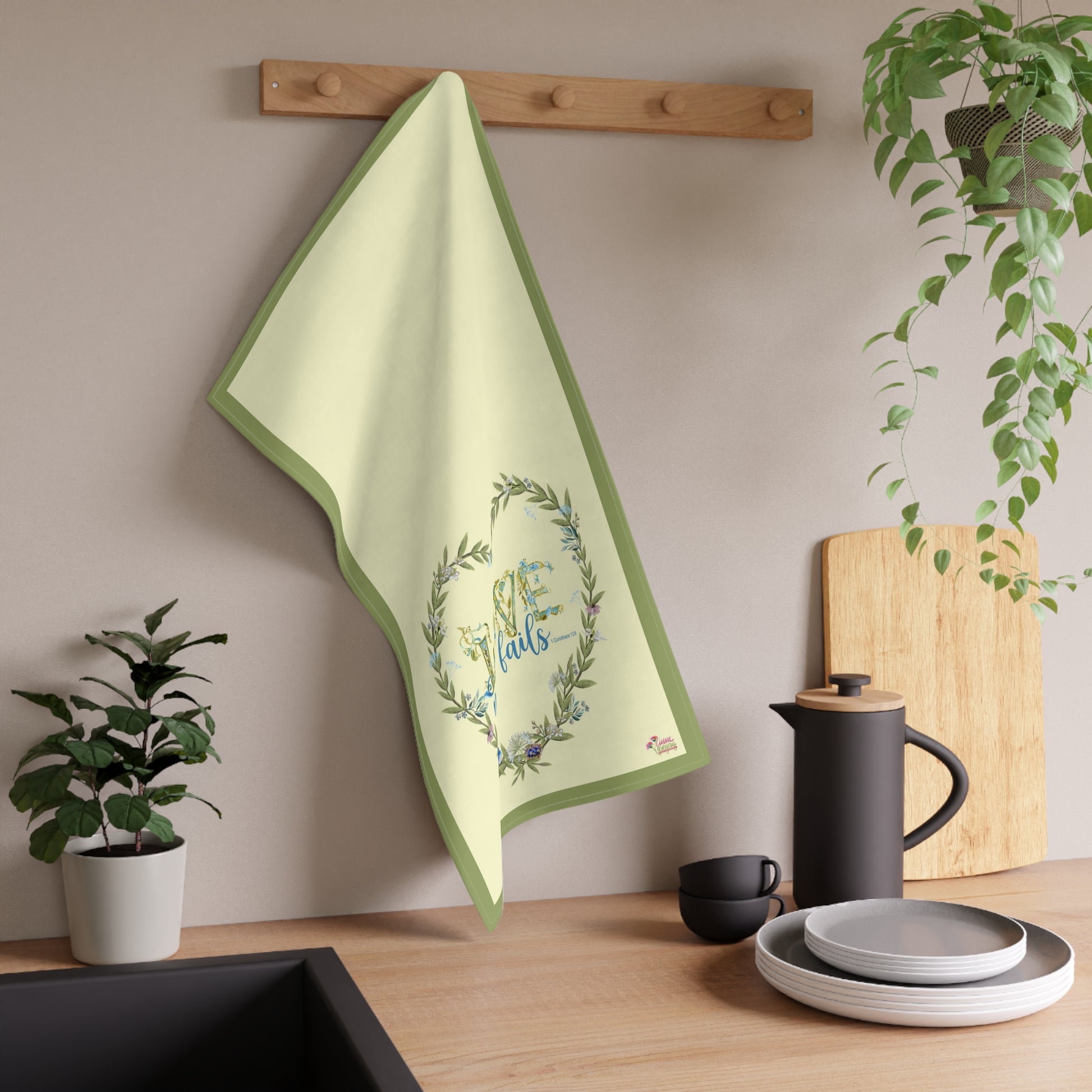 Tea towel with decorative design "Love Never Fails" hanging on a wooden rack in a kitchen setting.