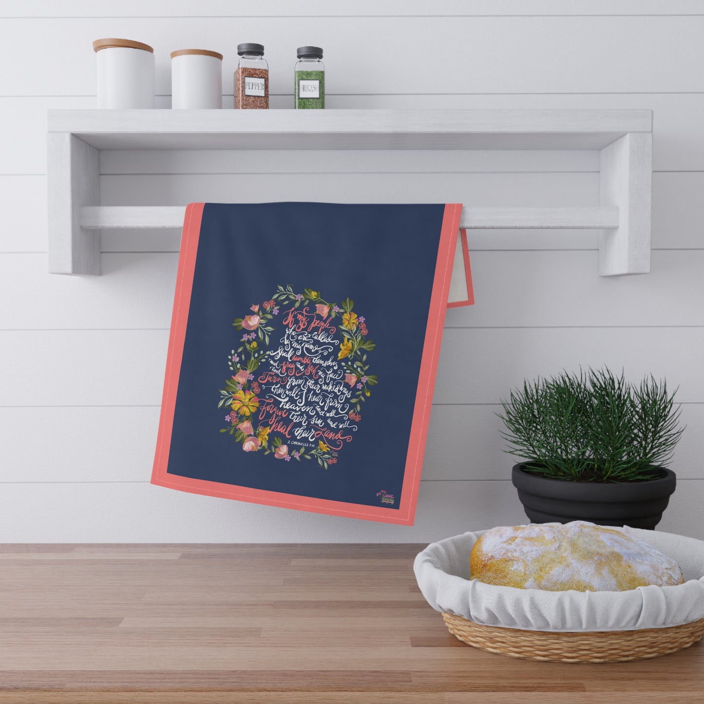 Navy blue kitchen tea towel with floral design and calligraphy Bible verse "If my People" hanging on a wooden shelf with a plant and basket below.