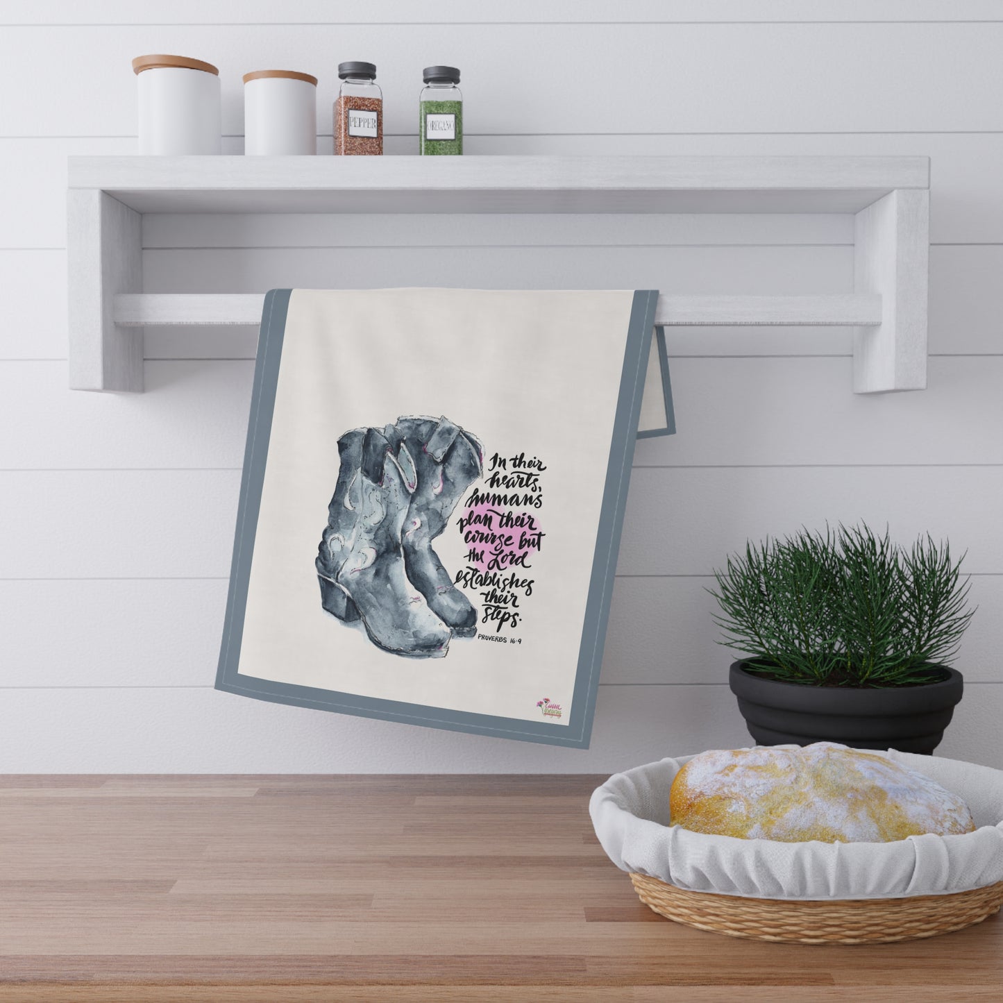 kitchen tea Towel with a design of cowboy boots and Bible verse "The Lord Establishes our Steps" hanging on a wall above a wooden surface with a plant and bread basket.