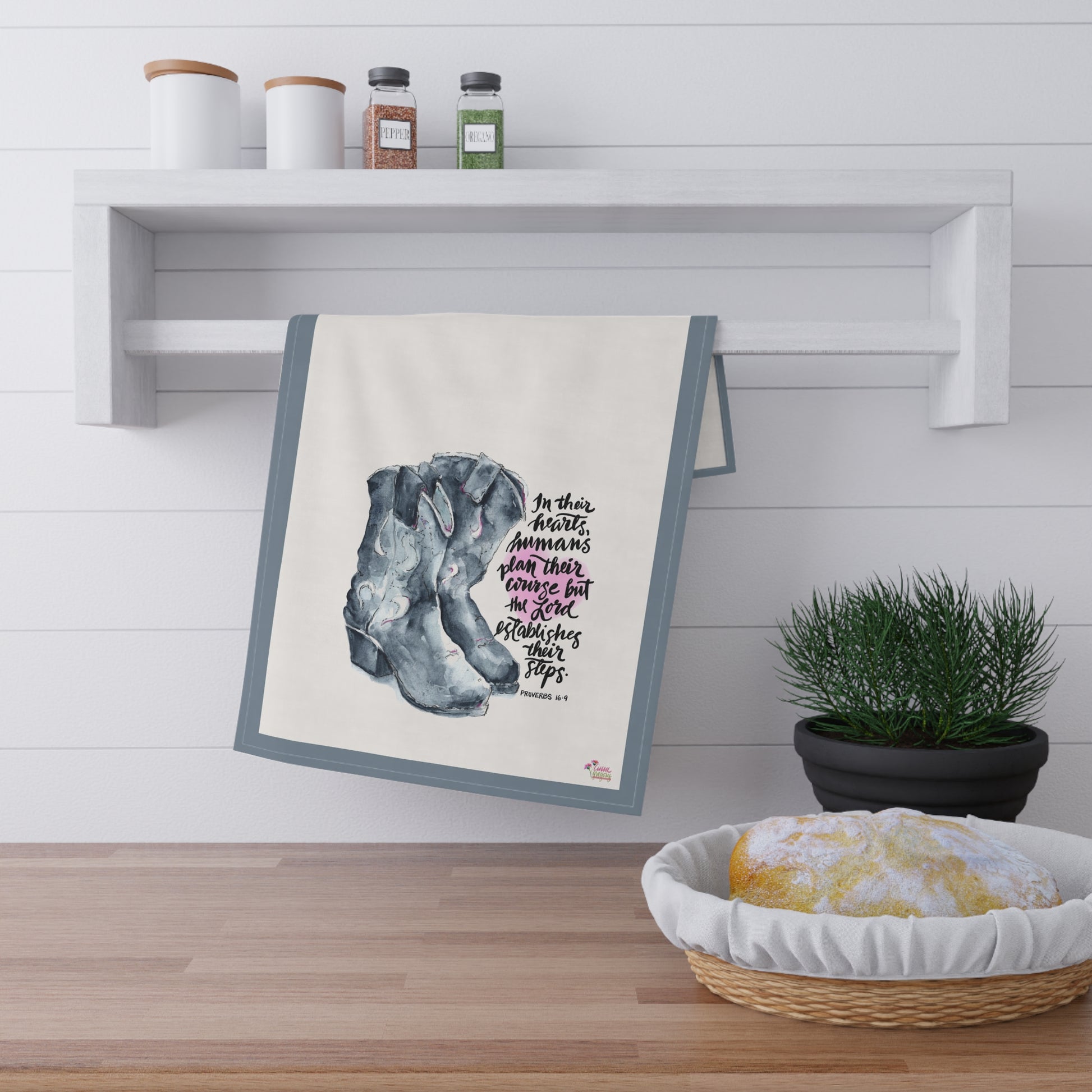 kitchen tea Towel with a design of cowboy boots and Bible verse "The Lord Establishes our Steps" hanging on a wall above a wooden surface with a plant and bread basket.