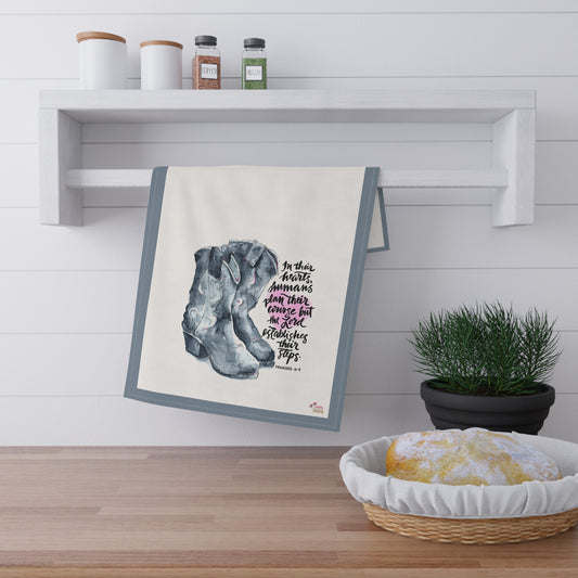 kitchen tea Towel with a design of cowboy boots and Bible verse "The Lord Establishes our Steps" hanging on a wall above a wooden surface with a plant and bread basket.