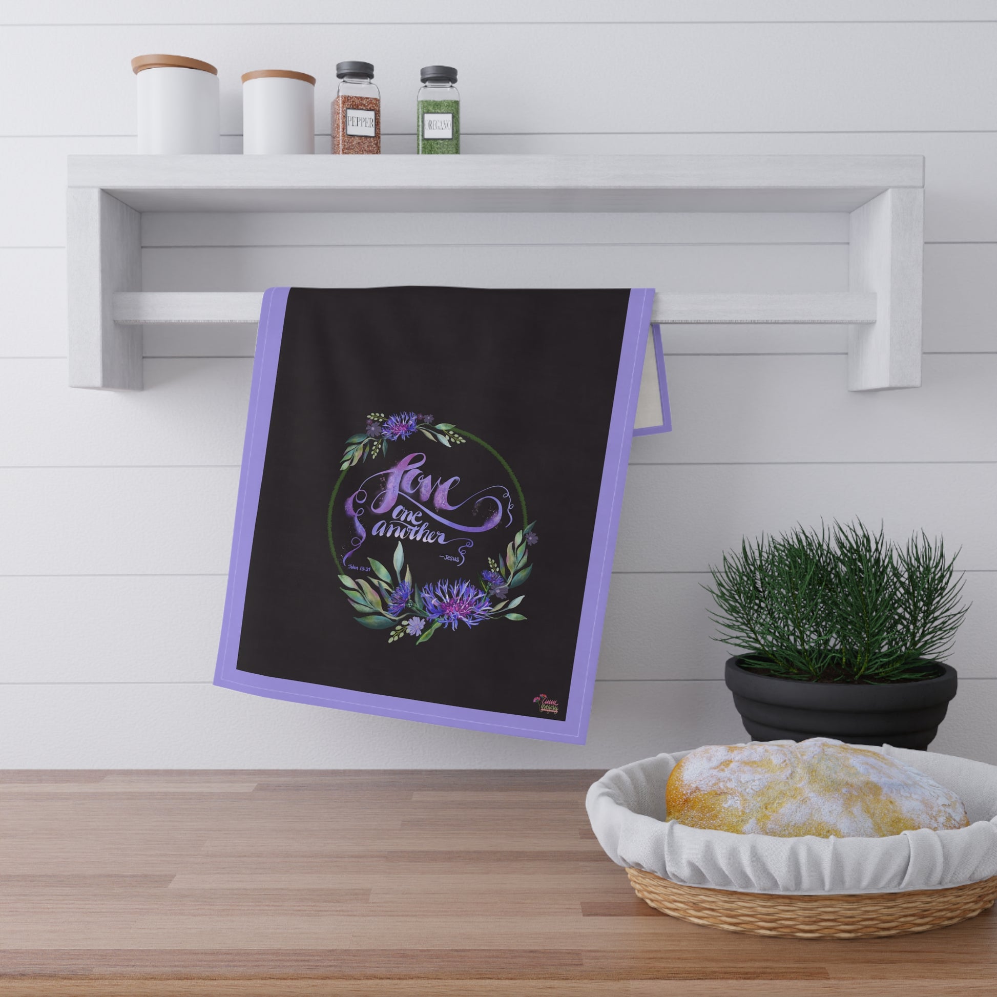 Black kitchen tea towel with purple border and floral design Bible verse "Love One Another" hanging on a wooden shelf with a plant and bread basket below.
