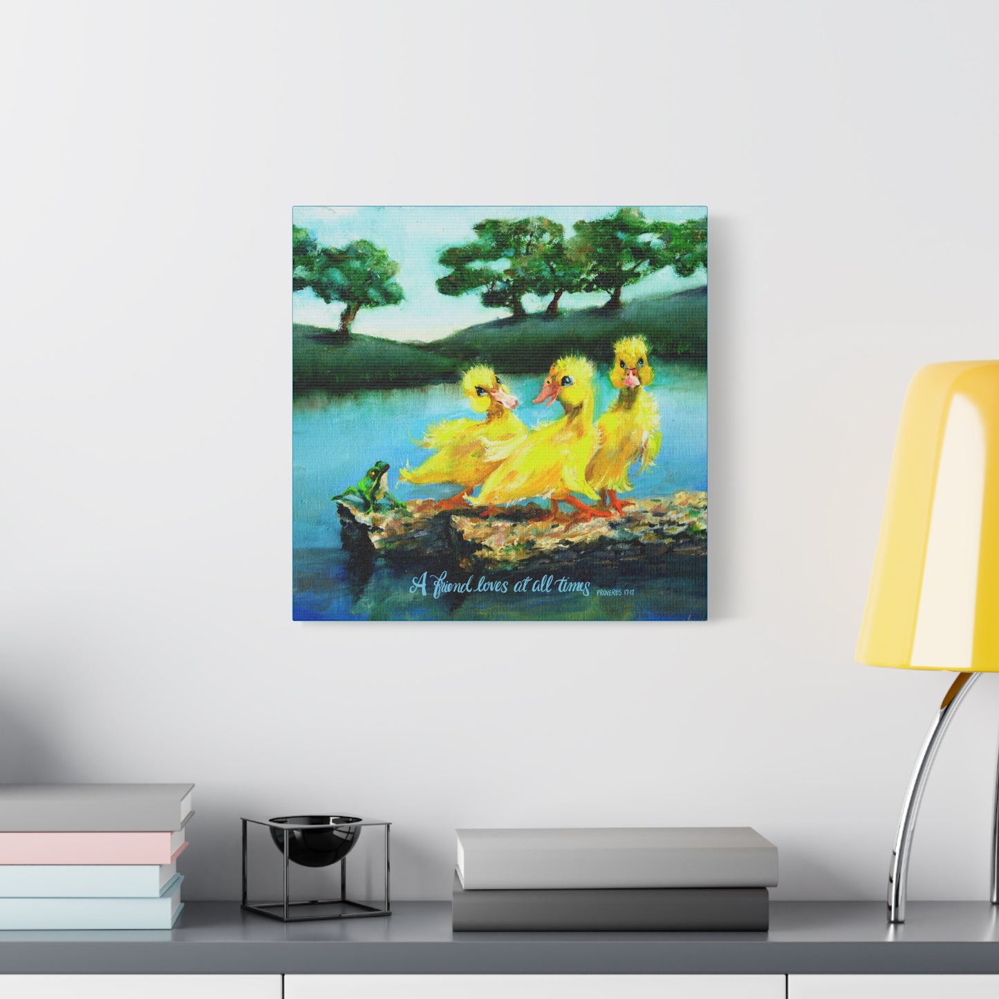 Canvas print of three yellow ducks on a log with trees and water in the background, displayed on a wall above a shelf. A Friend Loves at All Times.