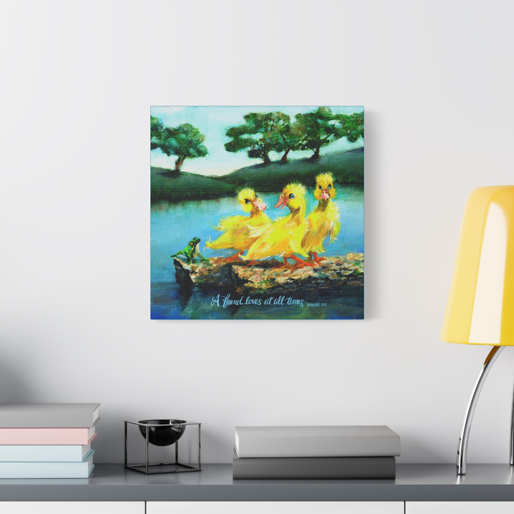 Canvas print of three yellow ducks on a log with trees and water in the background, displayed on a wall above a shelf. A Friend Loves at All Times.