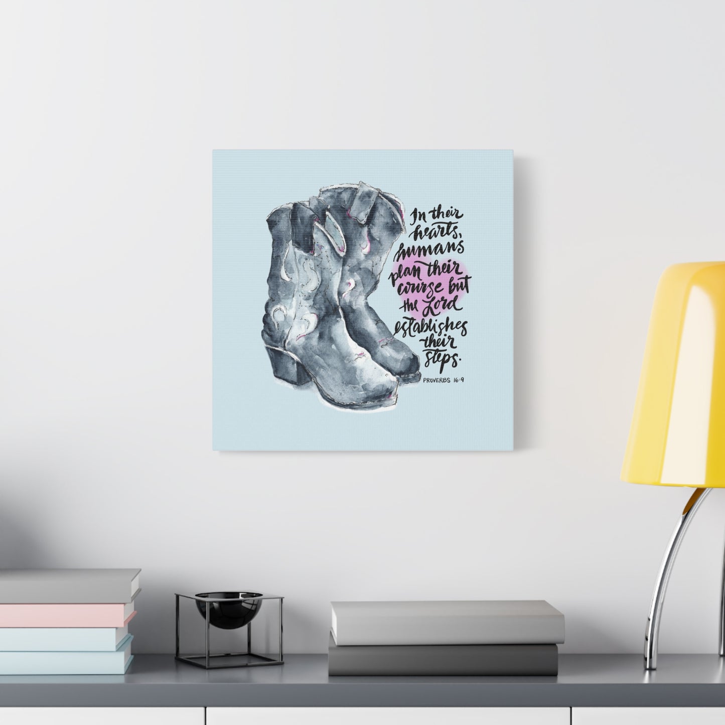 Canvas print with cowboy boots and Bible verse text, In their hearts humans plan their course but the Lord establishes their steps, on a light blue background, displayed on a shelf.