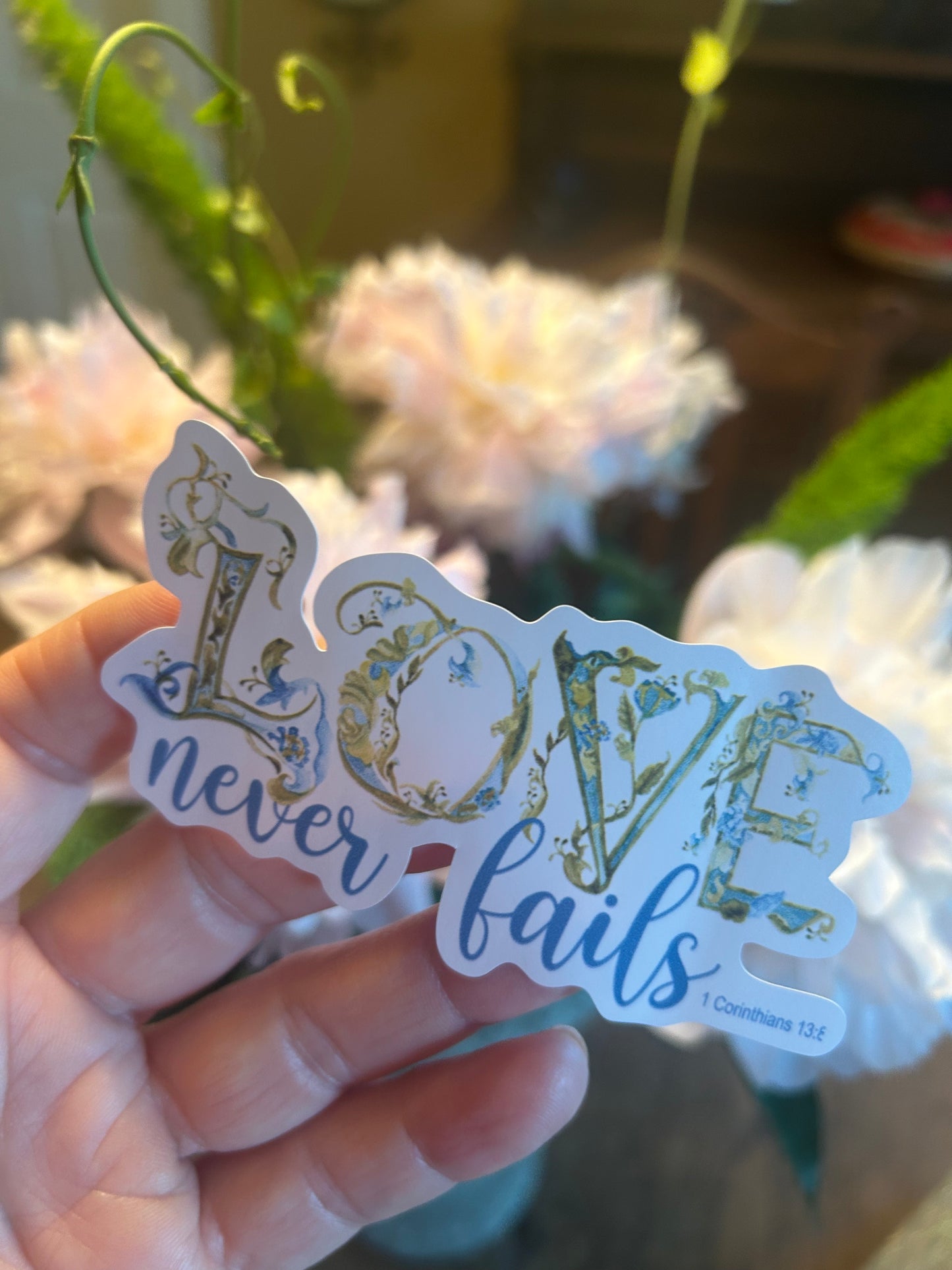 Hand holding a decorative sticker with 'Love never fails' text in front of flowers.