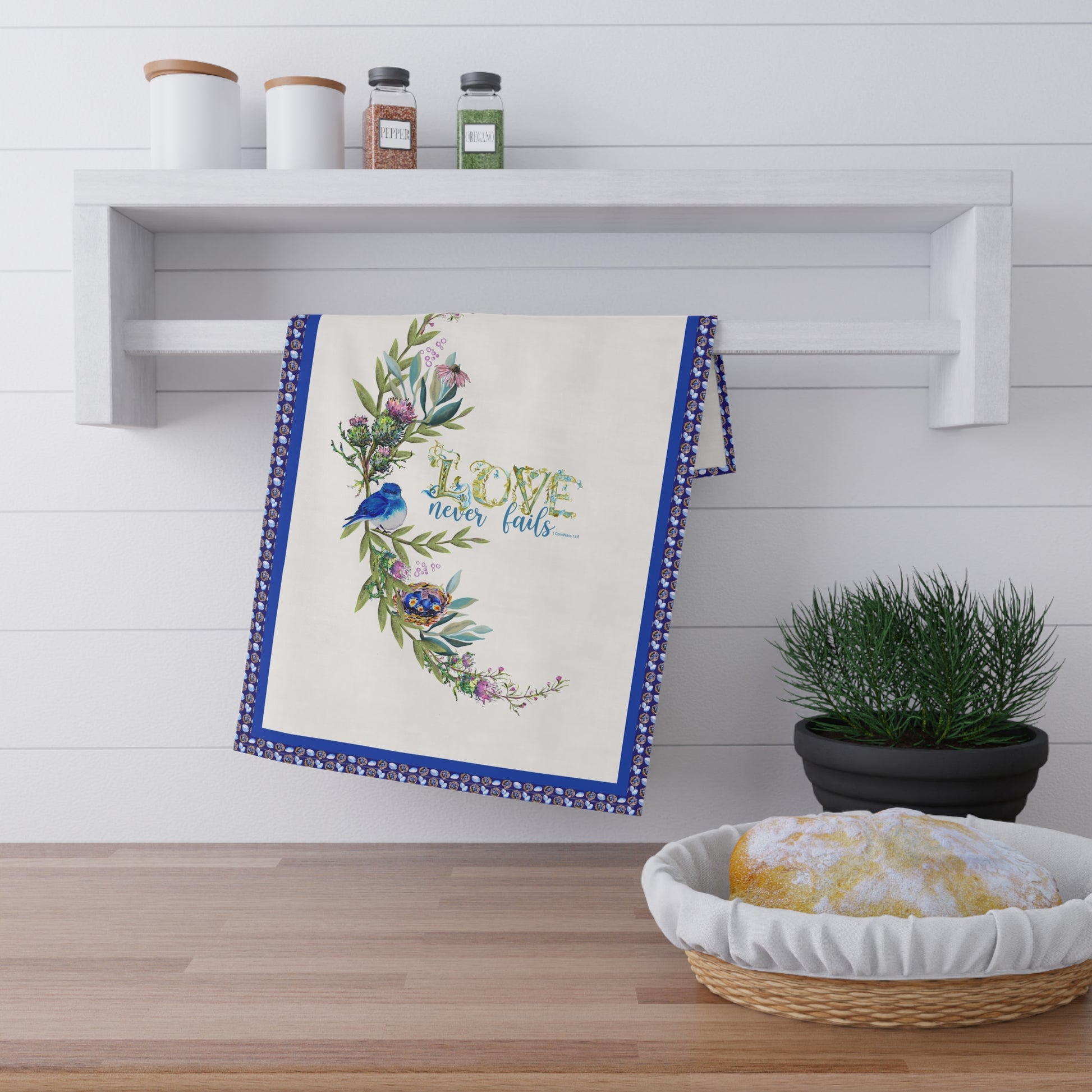 Decorative kitchen towel with floral design and Bible verse Love Never Fails, on a wooden surface with a plant and shelf in the background.