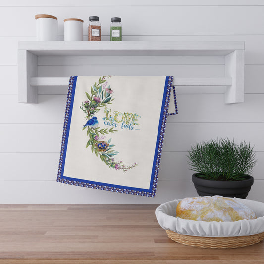 Decorative kitchen towel with floral design and Bible verse Love Never Fails, on a wooden surface with a plant and shelf in the background.