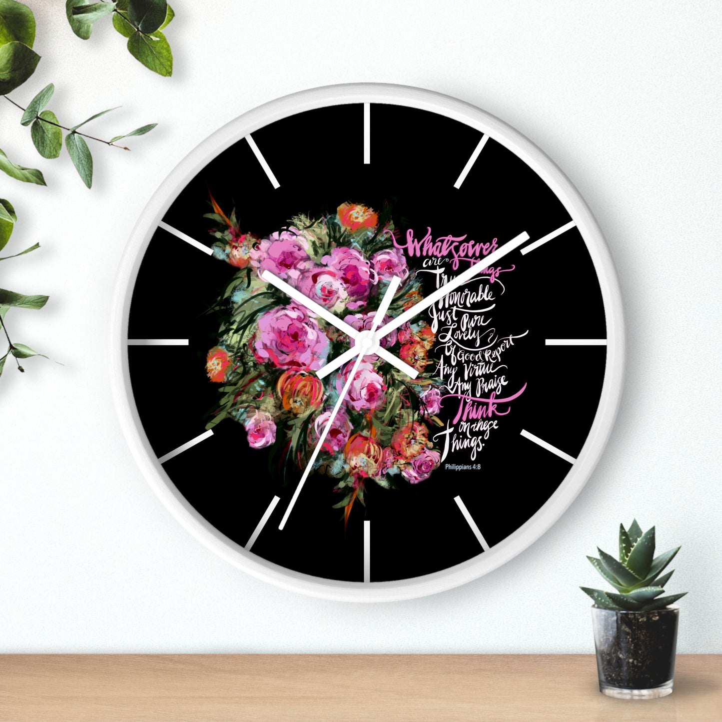 Wall clock with floral design and Bible verse text, Whatsoever things are true, honorable, just, Think on these Things, on a black background, and white frame, placed on a white surface with plants around.
