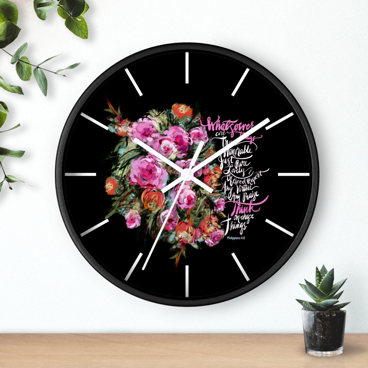 Wall clock on wall, with floral design and Bible verse text on a black background, placed on a wooden surface with plants.
